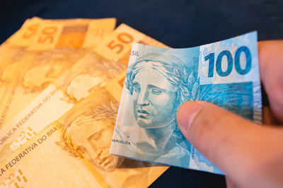 Foto em close-up de uma mão segurando uma nota de 100 reais sobre várias notas de 50 reais espalhadas.