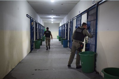 Foto mostra um corredor de uma prisão com celas dos dois lados com portões pintados de azul e dois agentes penitenciários fazendo a fiscalização