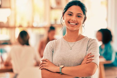 Foto: Canva Retrato de mulher feliz, de braços cruzados, com uma equipe de trabalho reunida ao fundo