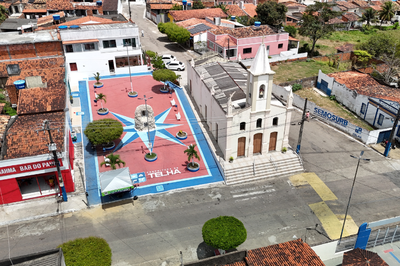Foto: Ascom Casa Civil Governo de Sergipe Vista aérea da praça central do município de Telha, Sergipe, destacando o piso colorido com uma rosa dos ventos e o letreiro da prefeitura. Ao lado, encontra-se uma igreja colonial branca com torre sineira, cercada por ruas pavimentadas e casas com telhados de barro.
