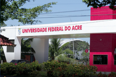 Foto: Divulgação Ufac Fachada da entrada da Universidade Federal do Acre (UFAC) com pórtico branco horizontal e letreiro em vermelho. À direita, destaca-se uma guarita alta revestida em pastilhas cor-de-rosa, cercada por vegetação e árvores.