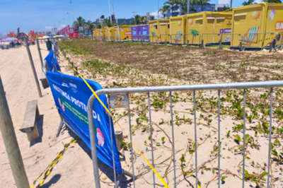 #PraTodosVerem: Foto colorida de uma área de restinga que foi pisoteada e está cercada por grades metálicas e faixas azuis de proteção, onde está escrito em destaque restinga protegida; ao fundo, veem-se containers amarelos.