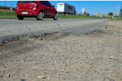 Fotografia em close do asfalto desgastado e com buracos em uma via marginal. Ao fundo, em plano desfocado, um carro vermelho e um caminhão trafegam sob céu azul.