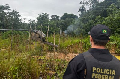 Costas de agente da Polícia Federal em frente a maquinário de garimpo improvisado no meio da floresta amazônica sob céu nublado.